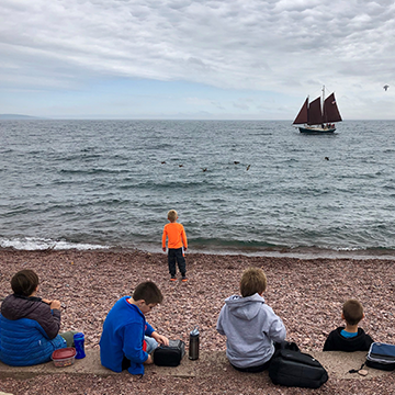 Students sitting on the beach as they watch a boat sail