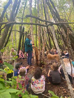 An adult talking to students and a dog inside a teepee log frame